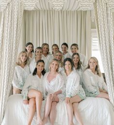 Bride in a lace robe and bridesmaids in matching white floral pajama shirts smiling together on a canopy bed in a bright bridal suite — wedding prep portrait.