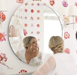Woman with blonde low bun checking her makeup in a round vanity mirror in a bright dressing room with pink floral wallpaper, wearing a sheer-sleeved white top.