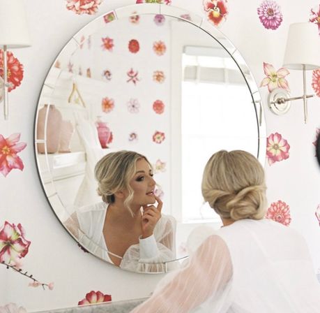 Woman with blonde low bun checking her makeup in a round vanity mirror in a bright dressing room with pink floral wallpaper, wearing a sheer-sleeved white top.