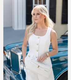 Chic blonde woman in a tailored white sleeveless outfit with gold buttons holding a white clutch, posing beside a teal vintage car on a sunlit street.