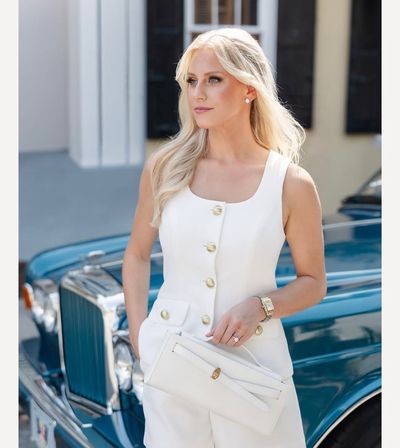 Chic blonde woman in a tailored white sleeveless outfit with gold buttons holding a white clutch, posing beside a teal vintage car on a sunlit street.