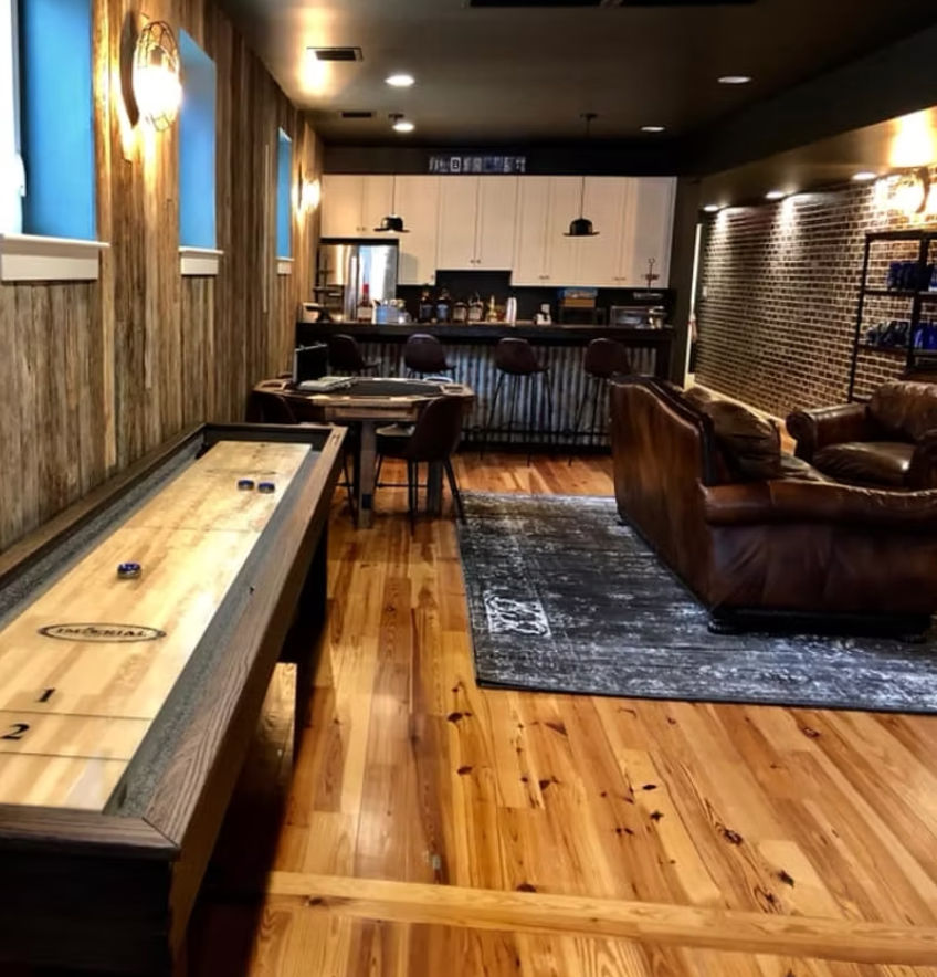 Cozy rustic basement game room with shuffleboard table, warm wood plank floors, leather sofa on area rug, and industrial-chic bar with pendant lights.