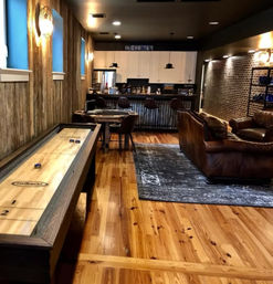 Cozy rustic basement game room with shuffleboard table, warm wood plank floors, leather sofa on area rug, and industrial-chic bar with pendant lights.