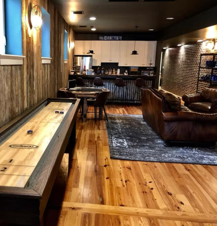Cozy rustic basement game room with shuffleboard table, warm wood plank floors, leather sofa on area rug, and industrial-chic bar with pendant lights.