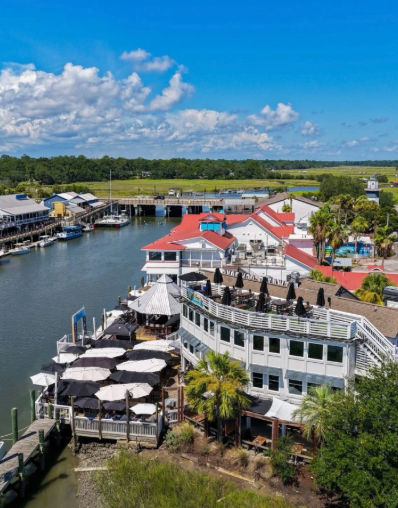 Aerial view of a coastal marina and waterfront dining with red-roofed buildings, umbrella-covered outdoor decks, docked boats, surrounding marshland and blue sky.