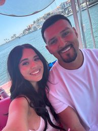 Two smiling people taking a selfie on a pink boat at a sunny marina, blue water with palm trees and waterfront homes in the background.