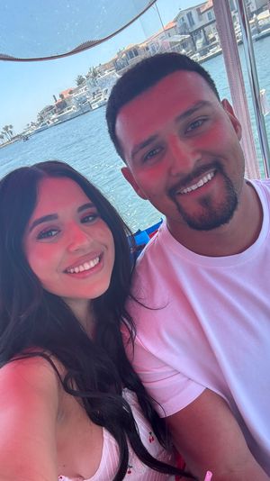 Two smiling people taking a selfie on a pink boat at a sunny marina, blue water with palm trees and waterfront homes in the background.