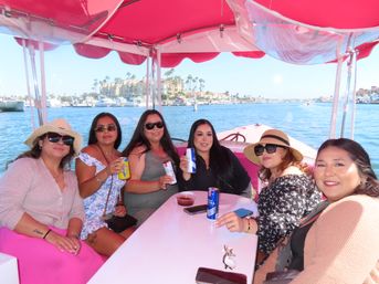 Group of friends on a pink‑canopied boat enjoying drinks in a sunny marina with palm trees, yachts and waterfront condos in the background.