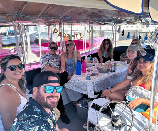 Group of friends enjoying a decorated pontoon boat party at a marina, banner overhead, table with snacks and drinks, sunglasses and hats on a sunny summer outing.