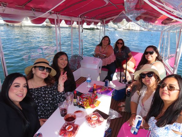 Smiling group of women on a pink‑canopied pontoon boat at a sunny marina with snacks and drinks on the table.