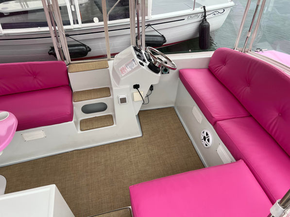Vibrant pink-cushioned pontoon boat interior with white helm and steering wheel, woven tan floor mat, stepped entry and wraparound bench seating docked at a marina.