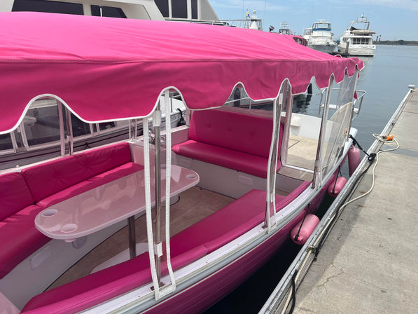 Bright pink canopy boat docked at a marina, with matching pink bench seating and table under a scalloped awning, moored beside a concrete dock with yachts in the background