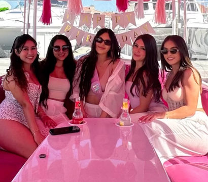 Five women in pink and white outfits smiling on a pink party boat at a marina, birthday banner and tassel decorations overhead and drinks on a glossy pink table.
