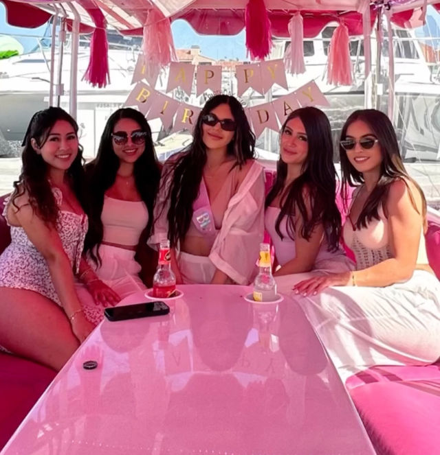 Five women in pink and white outfits smiling on a pink party boat at a marina, birthday banner and tassel decorations overhead and drinks on a glossy pink table.
