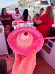Pink molded dessert topped with whipped cream and a cherry on a pink plate and spoon, held aboard a party boat with blurred friends and yachts at a sunny marina.