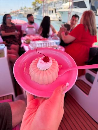 Pink molded dessert topped with whipped cream and a cherry on a pink plate and spoon, held aboard a party boat with blurred friends and yachts at a sunny marina.
