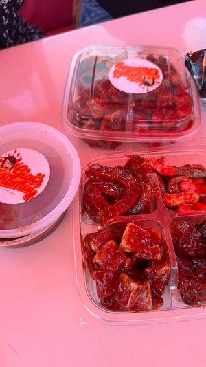 Plastic takeout containers of bright red, tangy chamoy-coated gummies, tamarind cubes and spicy candy pieces with a dipping sauce on a pink table