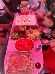 Pink-themed summer party spread: wooden charcuterie board with grapes, strawberries, raspberries, nuts, olives and crackers; bowl of cubed watermelon; tray of mini cherry-topped bundt cakes and a jar of pink punch.