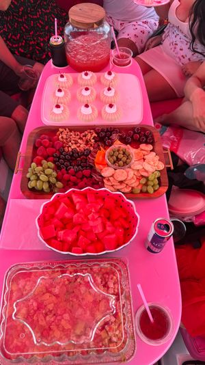 Pink-themed summer party spread: wooden charcuterie board with grapes, strawberries, raspberries, nuts, olives and crackers; bowl of cubed watermelon; tray of mini cherry-topped bundt cakes and a jar of pink punch.
