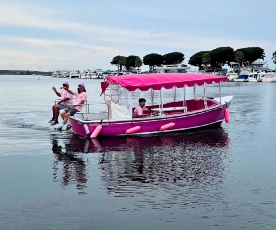 Bright pink canopy boat with crew in matching pink shirts cruising calm harbor waters past a yacht-lined marina under a pale blue sky.