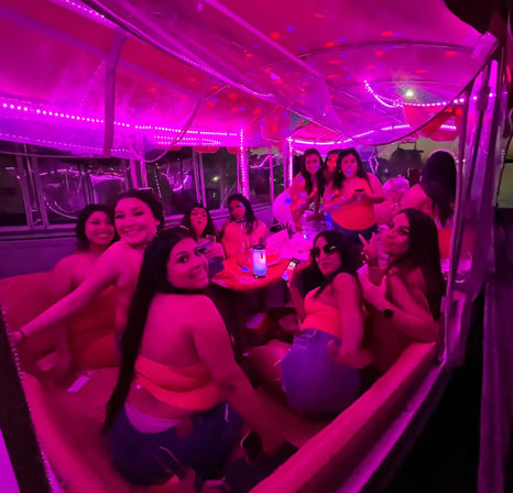 Group of friends partying inside a neon pink‑lit party bus at night, laughing around a table with drinks and LED string lights.