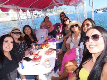 Group of women smiling for a selfie on a pink-canopied pontoon boat at a sunny marina, snacks and drinks on the table and yachts and waterfront buildings in the background.