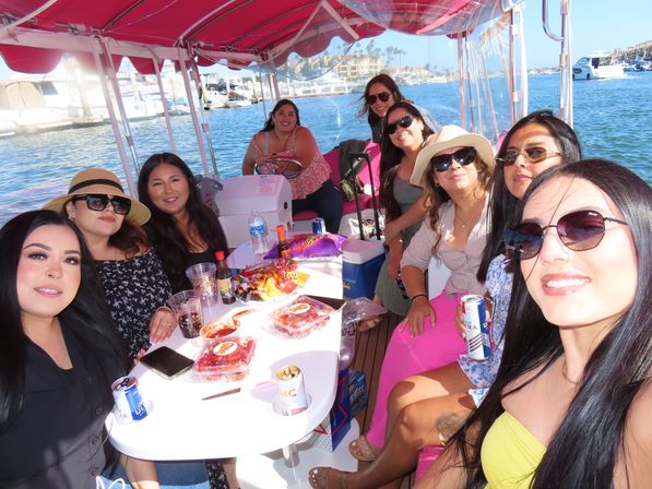Group of women smiling for a selfie on a pink-canopied pontoon boat at a sunny marina, snacks and drinks on the table and yachts and waterfront buildings in the background.