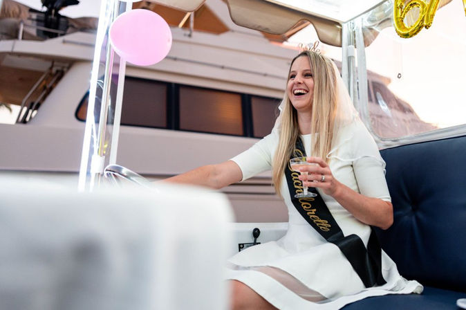 Smiling woman in white dress and veil wearing a bachelorette sash, holding champagne aboard a yacht with pink balloons