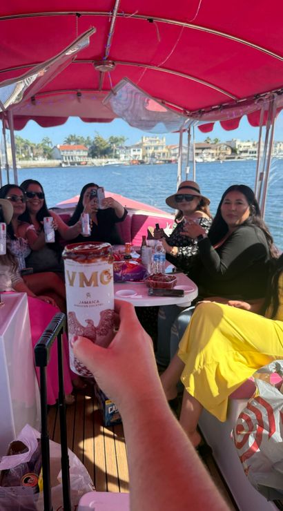 Friends toasting with canned drinks under a bright pink canopy on a party boat cruising a sunny waterfront lined with houses and palm trees