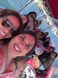 Selfie of a smiling group of friends on a pink-canopied boat at a sunny marina, with drinks on the table and yachts on calm blue water in the background.