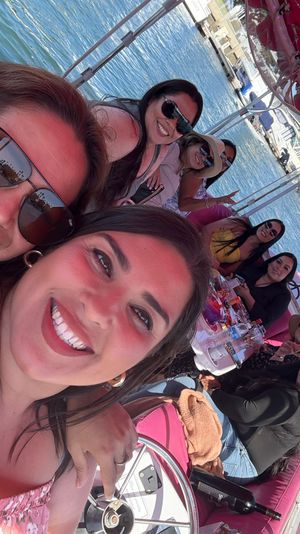 Selfie of a smiling group of friends on a pink-canopied boat at a sunny marina, with drinks on the table and yachts on calm blue water in the background.