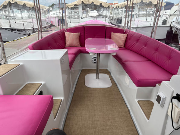 Bright pink cushioned U-shaped seating with matching pink table and cup holders on a pontoon boat interior at a marina dock, tan woven flooring and clear canopy panels visible