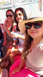 Three women in sunglasses and summer outfits, one wearing a wide-brimmed hat, enjoying canned drinks on a sunny boat ride with a marina and docked boats in the background.