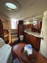 Cozy luxury yacht interior showing a compact galley and salon with glossy wood cabinets, curved countertop and sink, fridge, leather seating, oval skylight, steps to a sleeping cabin and a small table with blue-and-white flowers.
