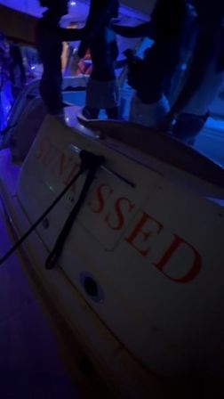 Nighttime yacht party on a boat deck — silhouetted people dancing under blue and purple lights, stern with partially visible red lettering.