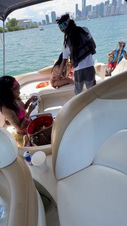 Group of friends on a motorboat near the Miami skyline, people in swimsuits lounging on white leather seats, man in a life vest by the bow, turquoise bay and high-rise cityscape in the background.