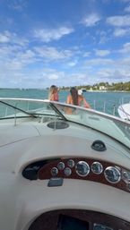 View from a yacht helm toward the bow with two people in swimsuits enjoying turquoise coastal waters, palm‑lined shoreline and a sunny blue sky