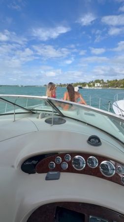 View from a yacht helm toward the bow with two people in swimsuits enjoying turquoise coastal waters, palm‑lined shoreline and a sunny blue sky