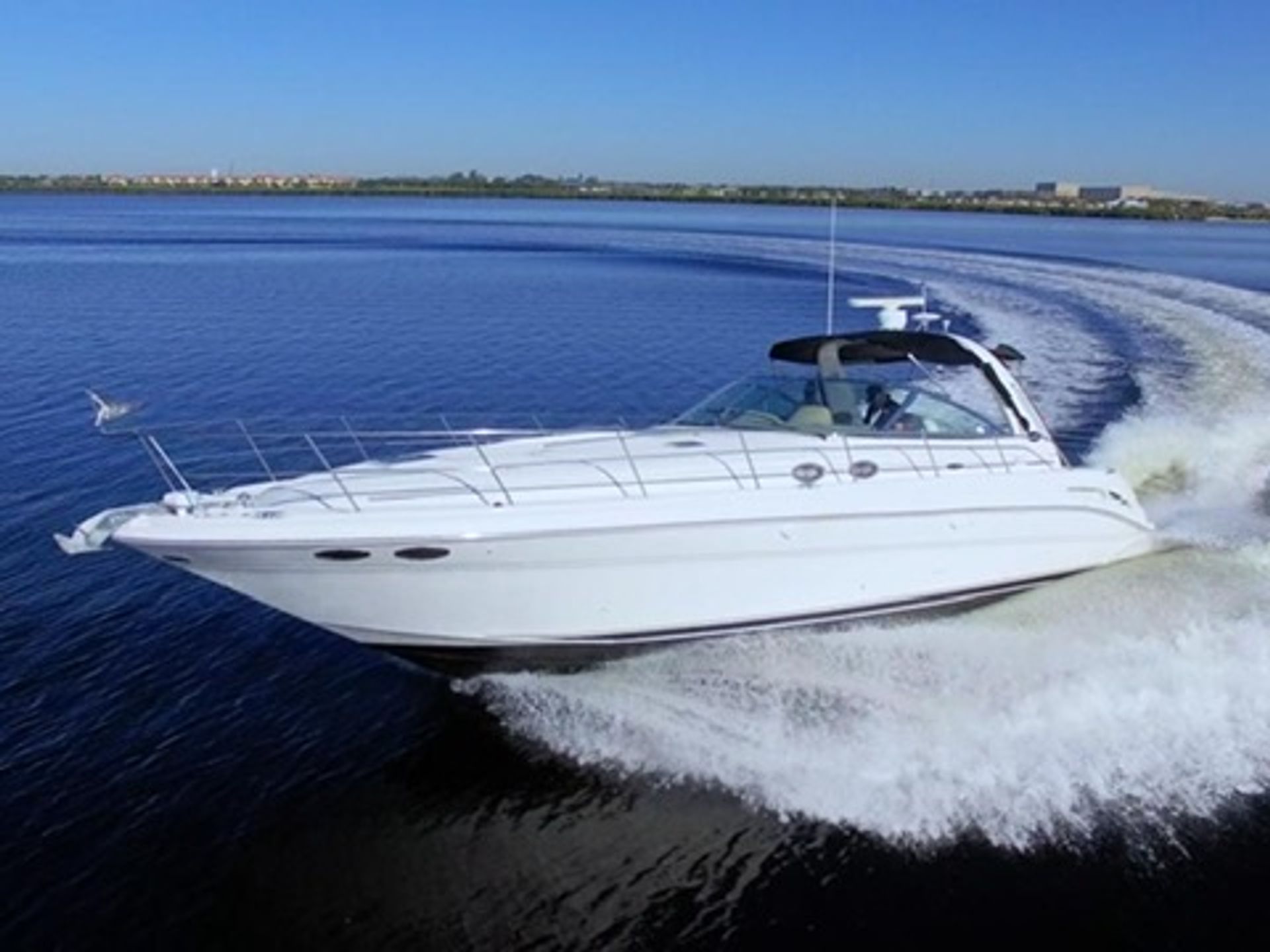 White luxury motor yacht speeding across calm blue water, cutting a curved wake with a faint shoreline on the horizon under a clear blue sky
