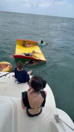 Group of adults relaxing on a bright yellow-and-red inflatable mat and colorful inner tubes beside a white boat swim platform in the open ocean under a cloudy sky