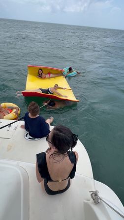 Group of adults relaxing on a bright yellow-and-red inflatable mat and colorful inner tubes beside a white boat swim platform in the open ocean under a cloudy sky