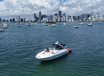 White motor yacht with people relaxing in the foreground on turquoise Biscayne Bay, dozens of anchored boats and the Miami skyline under a bright blue sky with fluffy clouds