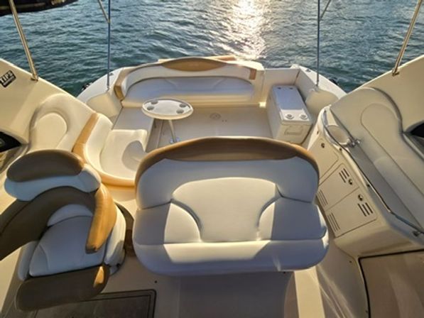 Motorboat cockpit with cream-and-tan cushioned bench seats, small round table and helm seating, overlooking shimmering water at sunset.