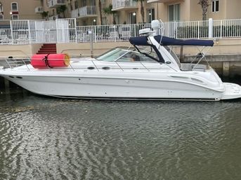 White cabin cruiser with navy bimini and red-yellow flotation fenders docked along a waterfront canal in front of beige condominium buildings with balconies and palm trees
