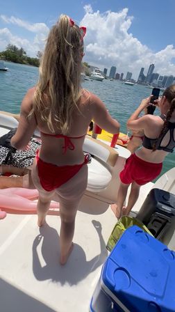 Sunny boating scene: two people in red swimwear on a boat deck, one taking a photo, with yachts and the Miami skyline under a blue sky.