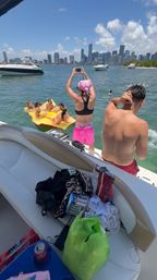 Friends enjoying a sunny boat day: a woman in pink shorts and a bandana snaps a photo of pals lounging on a bright yellow float in the water, with yachts and a distant city skyline under blue skies.