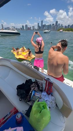 Friends enjoying a sunny boat day: a woman in pink shorts and a bandana snaps a photo of pals lounging on a bright yellow float in the water, with yachts and a distant city skyline under blue skies.