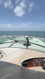 View from a yacht bow over calm turquoise ocean toward the horizon under a bright blue sky with scattered clouds, windshield and wood‑trimmed helm visible in foreground.