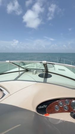View from a yacht bow over calm turquoise ocean toward the horizon under a bright blue sky with scattered clouds, windshield and wood‑trimmed helm visible in foreground.
