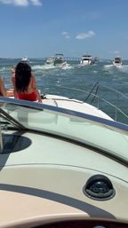 View from a motorboat bow with a woman in a red swimsuit sitting forward, a convoy of yachts cruising toward a distant coastal bridge under a sunny blue sky, deck and compass visible.